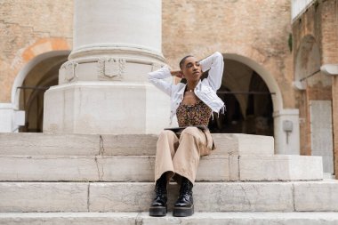 Short haired african american freelancer with laptop sitting on stairs on city street in Italy clipart