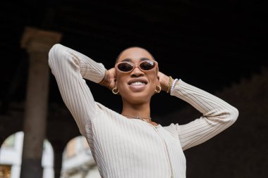 Gülümseyen, kısa saçlı, açık havada duran, güneş gözlüklü Afrikalı Amerikalı kadının alçak açılı görüntüsü. 