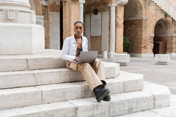 Short haired african american freelancer using laptop and looking at camera on stairs on urban street in Italy