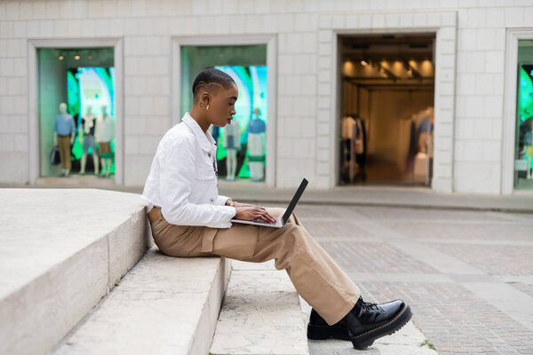 Side view of stylish african american freelancer using laptop with blank screen on stairs on urban street in Italy