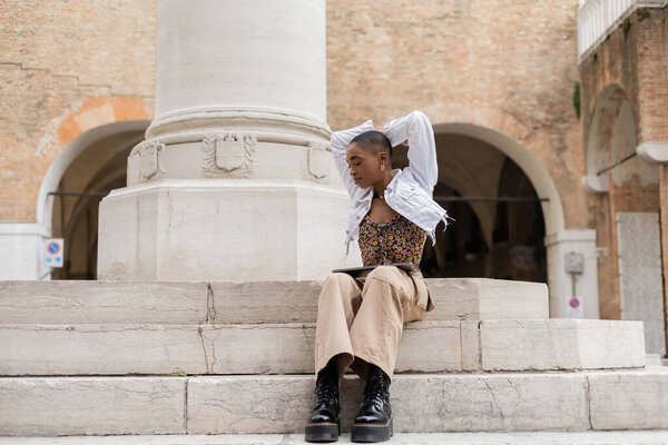 Trendy african american blogger with laptop sitting on stairs on urban street in Italy