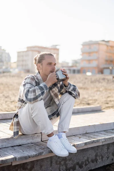 Long haired man holding cup while sitting on pier on beach in Italy — Stock Photo