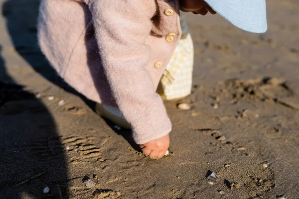 Vue recadrée du bébé en bas âge prenant coquille sur la plage de sable — Photo de stock