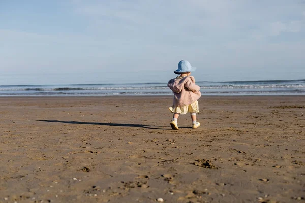 Vista posteriore della bimba in cappello panama passeggiando sulla spiaggia sabbiosa in Italia — Foto stock