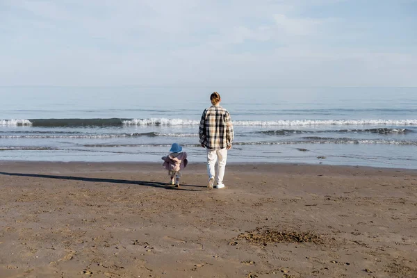 Back view of father and daughter in panama hat walking on beach near adriatic sea in Italy — Stock Photo