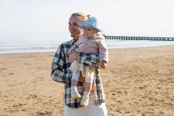 Positive father in shirt holding baby in panama hat on beach in Italy — Stock Photo