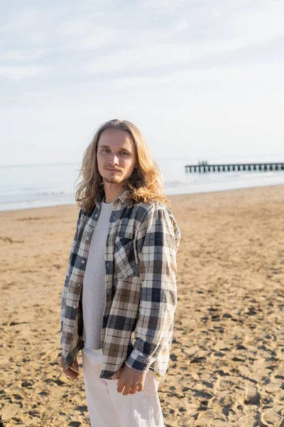 Hombre de pelo largo en camisa mirando a la cámara en la playa en Italia - foto de stock