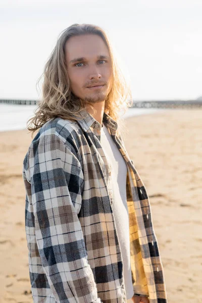 Ritratto di giovane uomo dai capelli lunghi che guarda la macchina fotografica sulla spiaggia in Italia — Foto stock