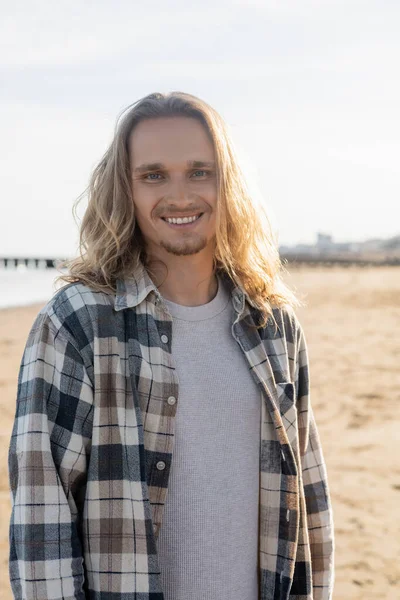 Porträt eines lächelnden langhaarigen Mannes, der am Strand in Italien in die Kamera blickt — Stockfoto