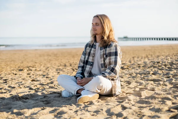 Молодой длинноволосый мужчина медитирует на пляже в Италии — стоковое фото