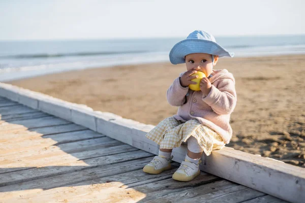 Niño comiendo manzana madura sentado en un muelle de madera en Italia - foto de stock