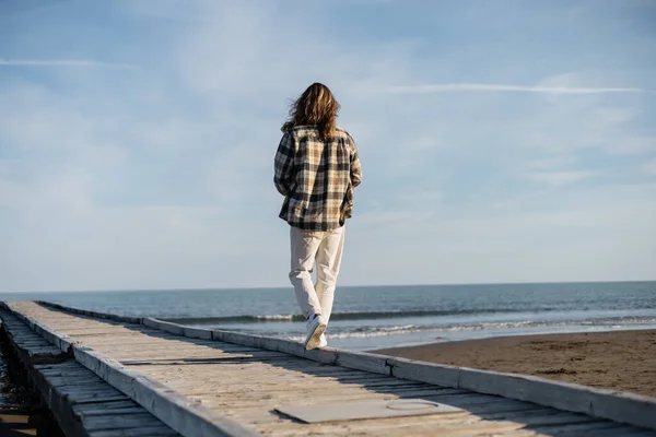 Vista posteriore dell'uomo dai capelli lunghi che cammina sul molo di legno vicino al mare Adriatico in Italia — Foto stock