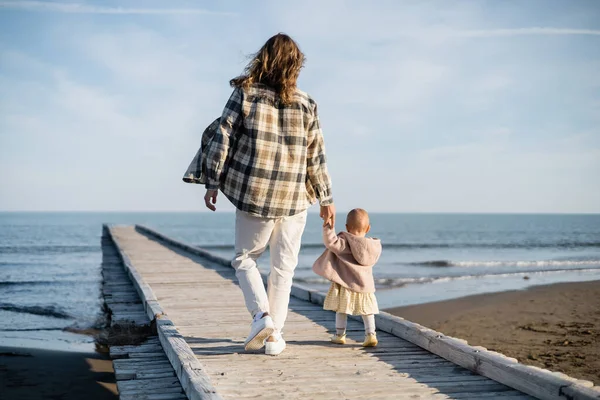Vista posteriore di uomo dai capelli lunghi e bambina che cammina sul molo di legno vicino al mare Adriatico in Italia — Foto stock