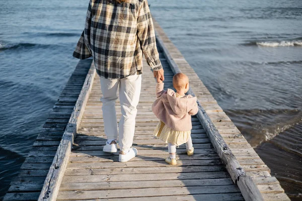 Вид сзади на родителя и девочку, прогуливающихся по деревянному пиру над Адриатическим морем в Тревизо — стоковое фото