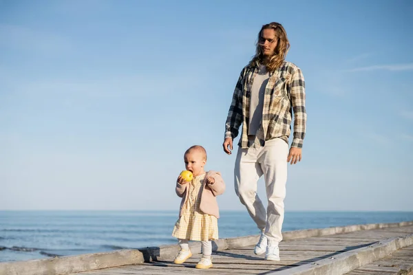 Young man walking near baby daughter with apple on pier on beach in Italy — Stock Photo