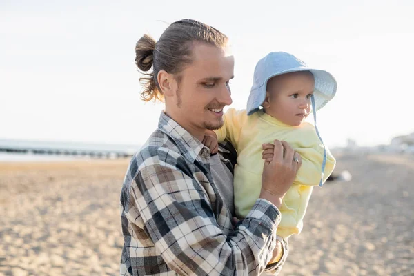 Papa souriant tenant bébé en chapeau panama sur la plage de Trévise — Photo de stock