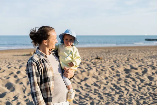 Positive parent holding baby in panama hat on beach near sea in Italy — Stock Photo