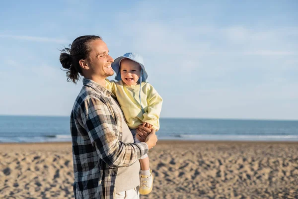 Langhaariger Vater hält fröhliche Tochter am Strand am Meer in Italien — Stockfoto