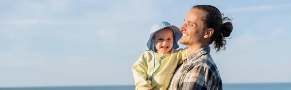 Dad in shirt holding toddler child in panama hat with sea at background, banner — Stock Photo