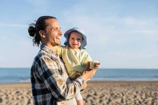 Positive father holding smiling child in panama hat on beach near sea in Italy — Stock Photo