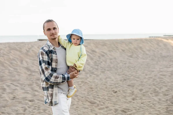 Молодой человек в рубашке держит дочь в панамской шляпе на пляже — стоковое фото