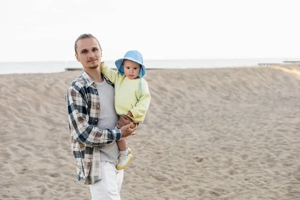 Young man in shirt holding daughter in panama hat on beach — Stock Photo