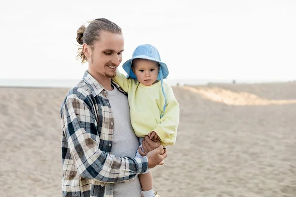 Длинноволосый мужчина в рубашке держит ребенка на пляже — стоковое фото