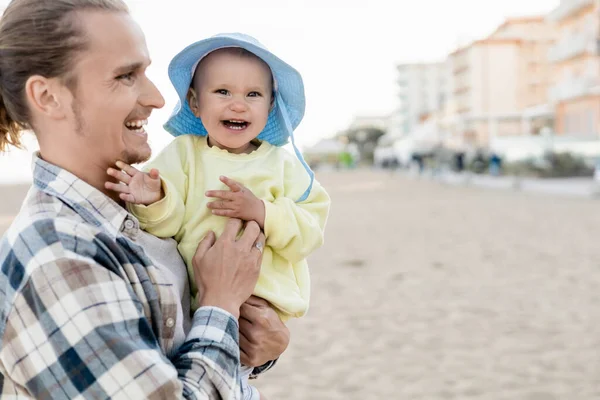 Positive man holding baby girl in panama hat on beach in Treviso — Stock Photo