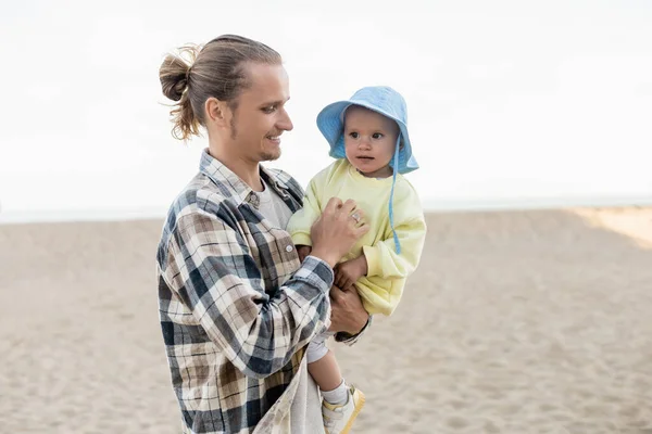 Papa aux cheveux longs tenant bébé en chapeau panama sur la plage — Photo de stock