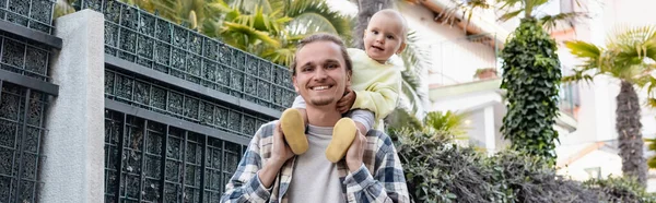 Jovem segurando bebê nos ombros e sorrindo para a câmera na rua urbana em Treviso, banner — Fotografia de Stock