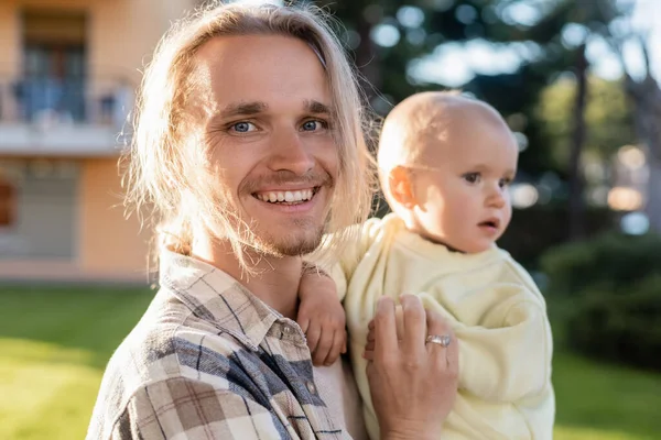 Positive man holding blurred baby and looking at camera outdoors — Stock Photo