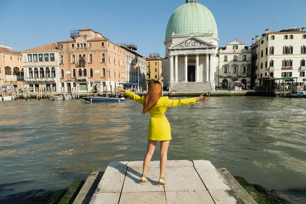 Visão traseira da mulher em roupas amarelas de pé com as mãos estendidas no cais do Grande Canal veneziano — Fotografia de Stock