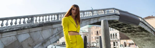 Руда жінка в жовтому модному костюмі позує біля венеціанського мосту на фоні, банер — стокове фото