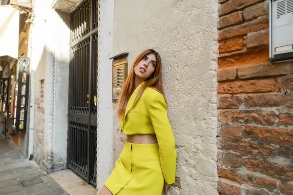 Модная женщина с рыжими волосами смотрит в сторону, позируя рядом с венецианским зданием — стоковое фото