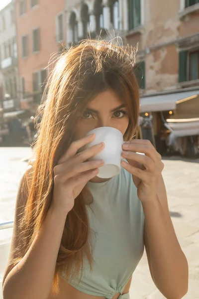 Redhead woman looking at camera while drinking coffee on blurred street in Venice — Stock Photo