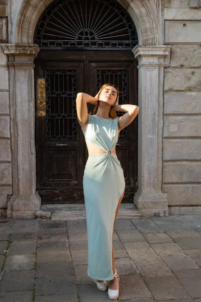 Mulher em vestido elegante posando com os olhos fechados e as mãos atrás da cabeça na rua veneziana — Fotografia de Stock