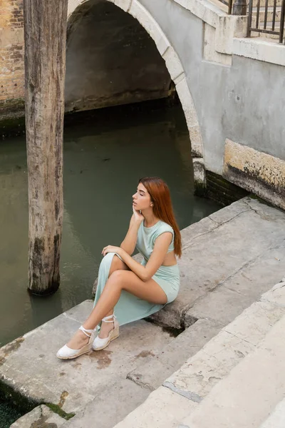 Високий кут зору стильна жінка з рудим волоссям, що сидить на сходах біля міського венеціанського каналу — стокове фото