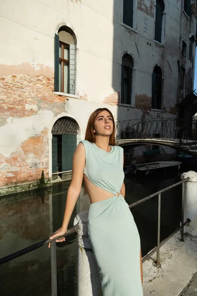 Руда жінка в елегантній сукні, що спирається на паркан біля каналу на венеціанській вулиці — стокове фото