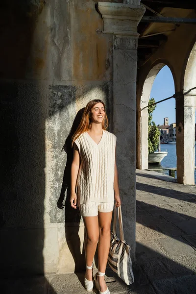 Longitud completa de la mujer feliz con los ojos cerrados de pie con el bolso rayado cerca de la pared de piedra en Venecia - foto de stock