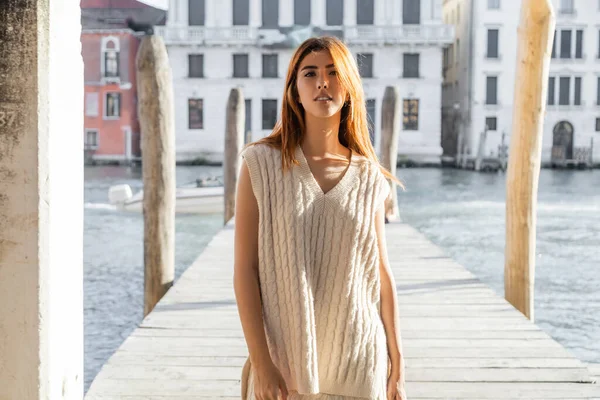 Junge Frau im ärmellosen Pullover blickt in die Kamera in der Nähe der verschwommenen Seebrücke in Venedig — Stockfoto