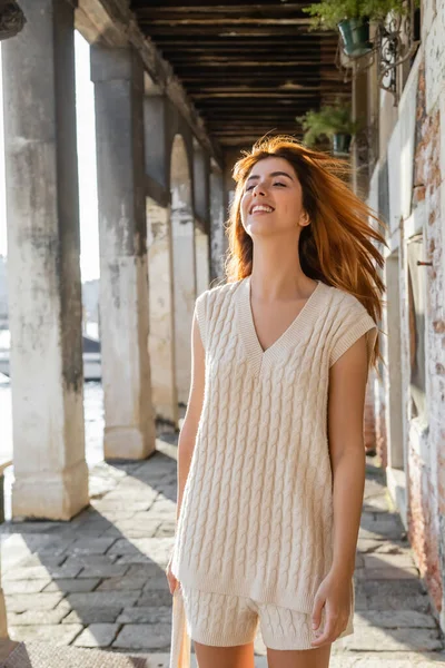 Mujer pelirroja feliz en jersey sin mangas de pie en el viento en la calle veneciana - foto de stock