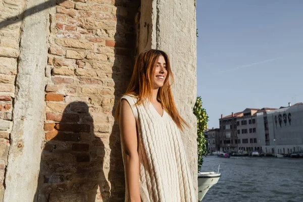 Cheerful redhead woman in sleeveless jumper smiling near stone wall in Venice — Stock Photo