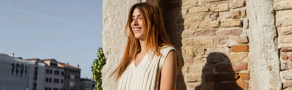 Mujer joven con el pelo rojo sonriendo cerca de la pared de piedra en Venecia, pancarta - foto de stock