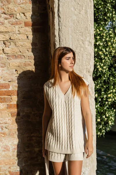 Mujer con estilo en jersey sin mangas mirando lejos cerca de la pared de piedra en Venecia - foto de stock
