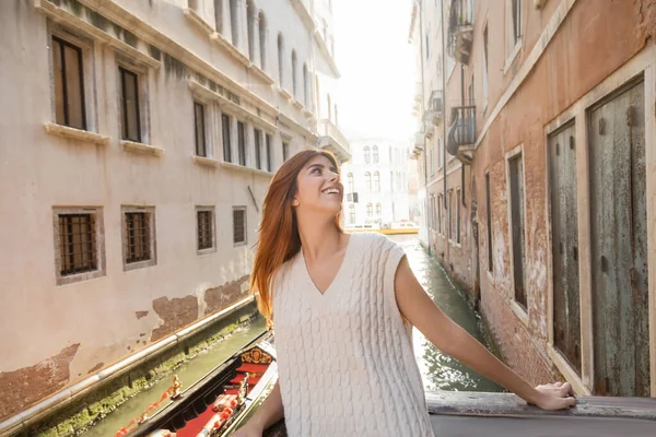 Mujer pelirroja feliz en prendas de punto de verano mirando edificios medievales en Venecia - foto de stock