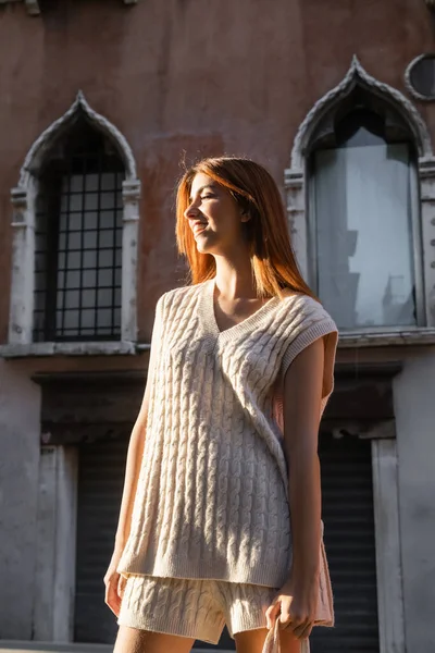 Молодая женщина в свитере без рукавов улыбается возле размытого здания в Венеции — стоковое фото