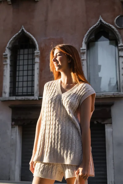 Giovane donna in maglione senza maniche sorridente vicino edificio sfocato a Venezia — Foto stock