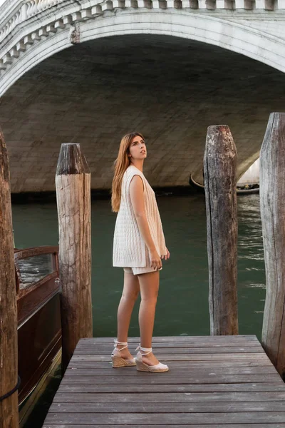 Mujer delgada en ropa de punto de verano y sandalias de cuña mirando hacia otro lado en el muelle de madera en Venecia - foto de stock