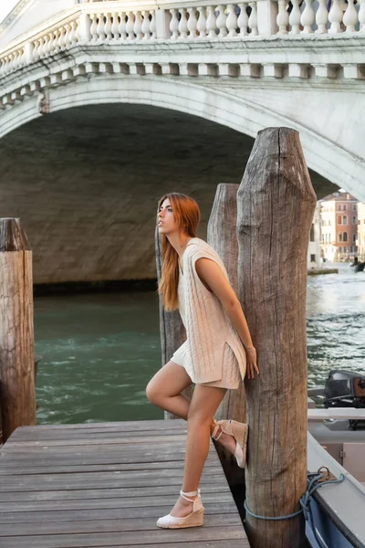 Стильная женщина в клиновых сандалиях, опирающаяся на деревянные сваи возле моста через Гранд-канал в Венеции — стоковое фото