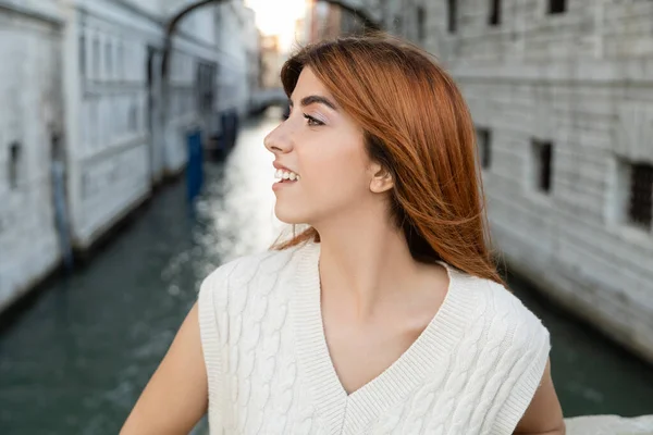 Mujer joven y feliz con el pelo rojo mirando hacia otro lado sobre fondo borroso en Venecia - foto de stock